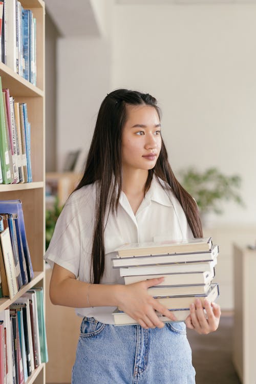 Free A Woman in White Polo Shirt Standing while Holding a Stack of Books Stock Photo