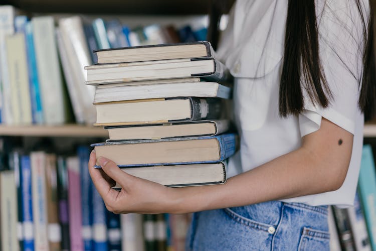 A Person In White Shirt Carrying A Stack Of Books