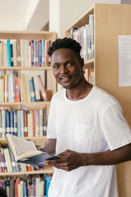 A Man in White Shirt Leaning on the Shelf while Holding a Book