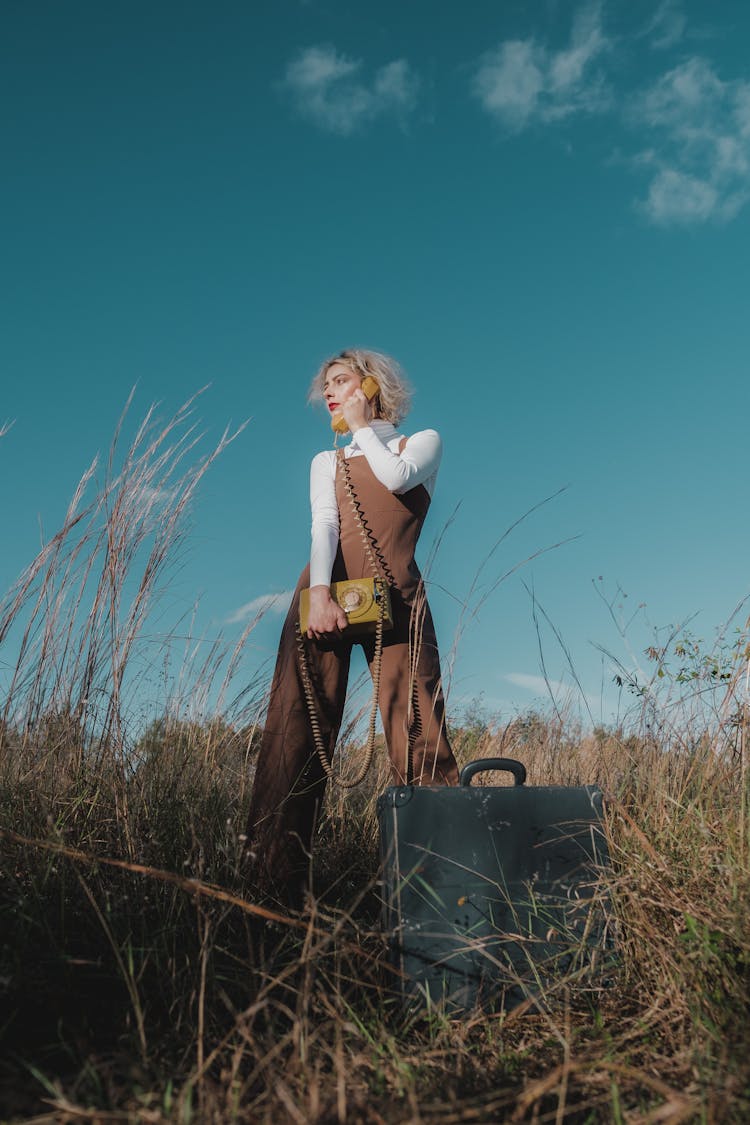 A Woman In White Long Sleeves And Brown Jumper Standing On Grass Field While Holding A Telephone