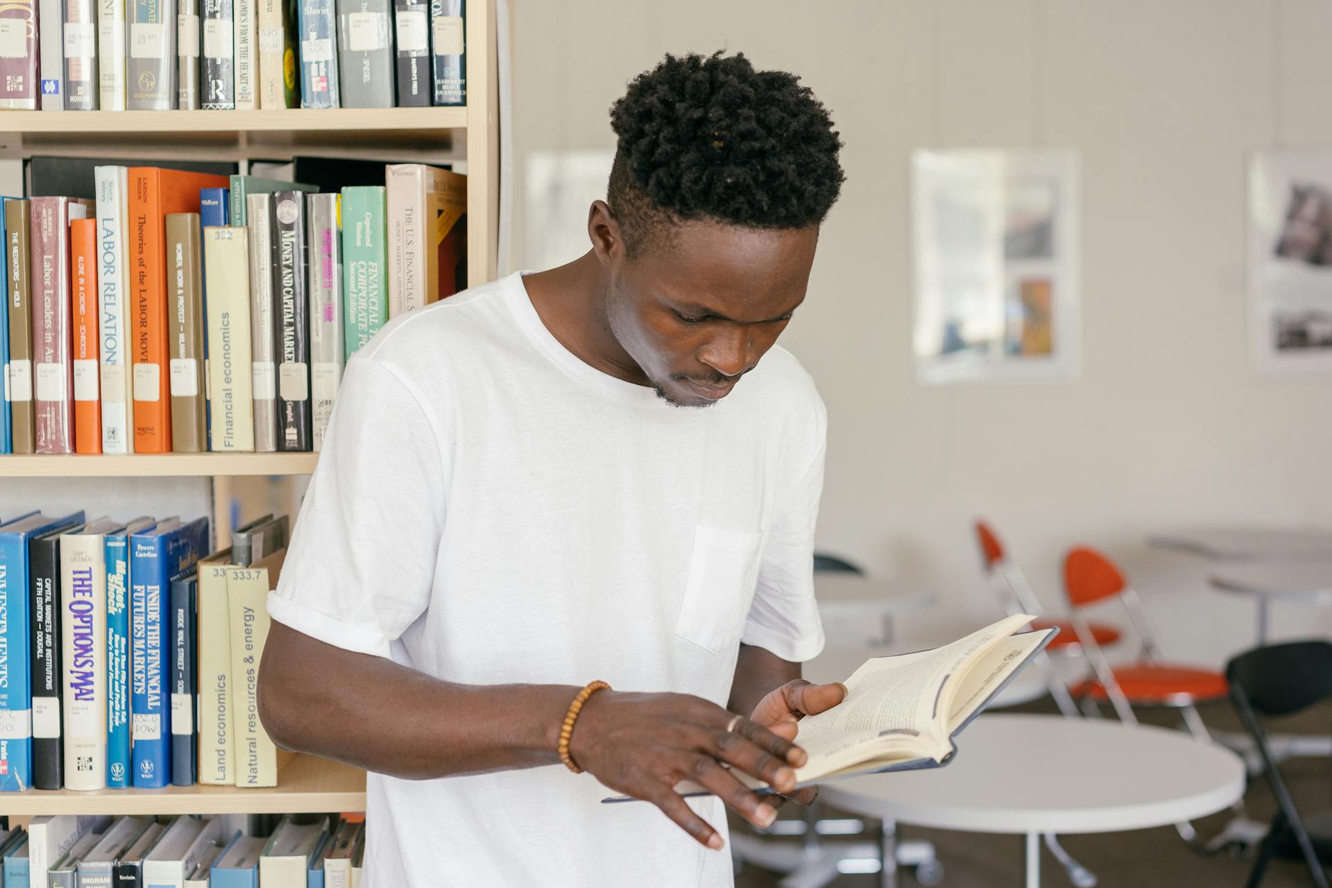 A young man in a white shirt reading a book in a modern library setting.