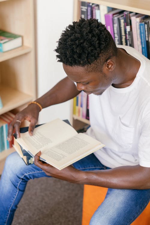 A Man Holding a Book While Sitting Near the Bookshelves