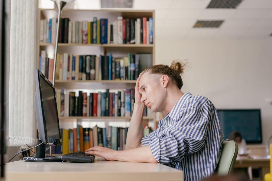A young student working on a computer in a library setting, deep in thought.