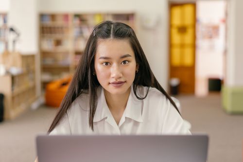 Free Asian woman using a laptop in a modern library setting, focused and engaged. Stock Photo