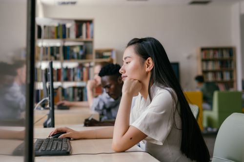 Free Asian woman studying diligently on a computer in a vibrant university library setting. Stock Photo