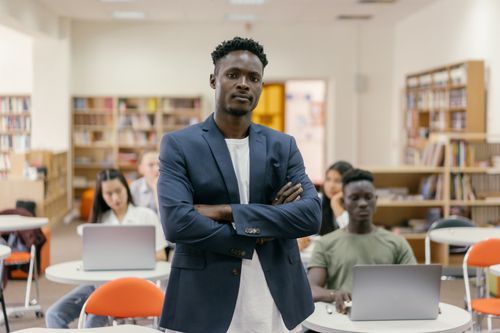 Free Teacher standing confidently in a library classroom with diverse students using laptops. Stock Photo