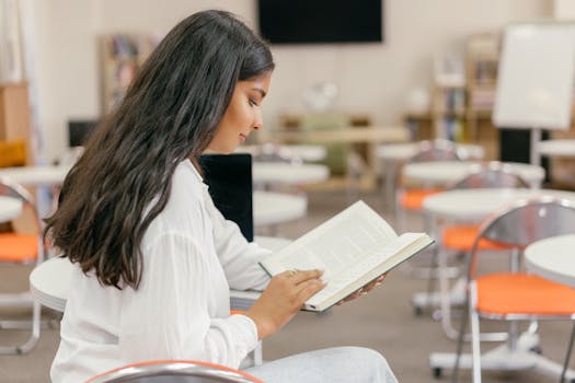 Young woman engrossed in reading a textbook in a modern, empty classroom setting.
