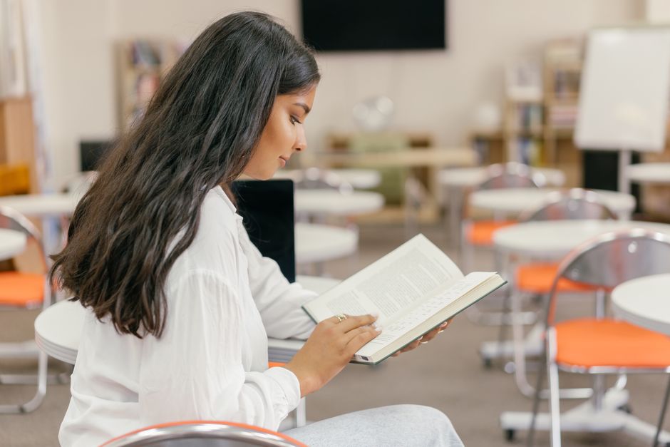 Young woman engrossed in reading a textbook in a modern, empty classroom setting.