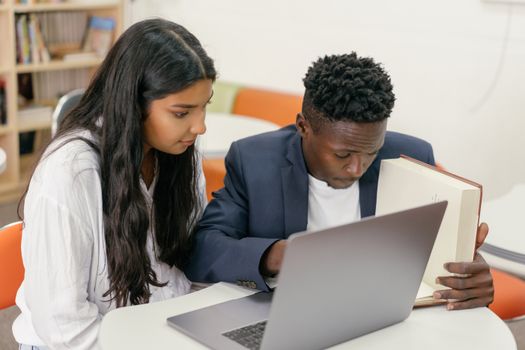 Two students collaborating with a book and laptop in a modern library environment.