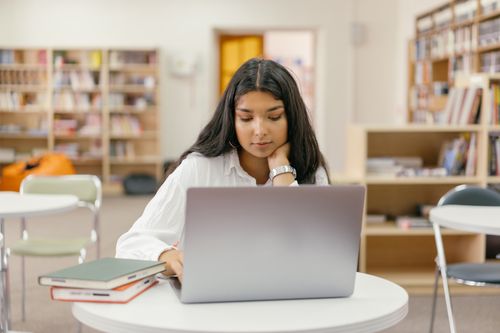 Free A young woman in a library studying online with a laptop and books around her. Stock Photo