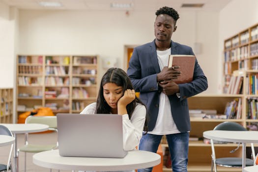 A student studies on a laptop while a teacher supervises in a university library.
