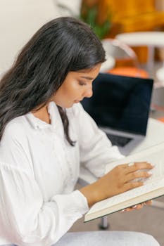 A teenager intently reading a book indoors, with a laptop nearby, creating an educational atmosphere.