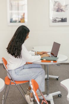 Side view of a young woman working on a laptop at a desk in a modern office setting, wearing casual attire.