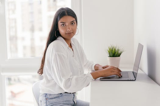 Young woman typing on a laptop at a white desk in a bright, modern room.