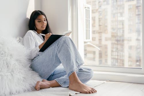 Free A Woman Sitting Near the Glass Window while Writing on the Book Stock Photo