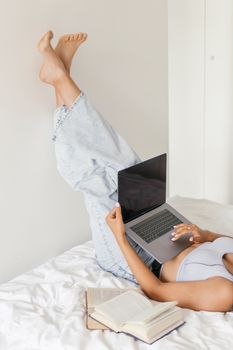 Adult woman lying on bed with legs up, using a laptop for study with books around.