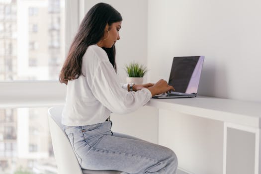 Young woman in a white shirt working on a laptop at a minimalist home office desk.