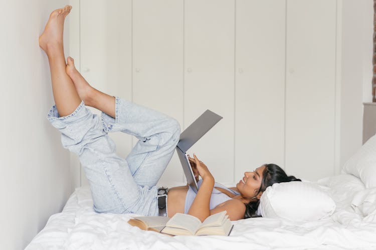 A Woman Lying On Bed With Legs Up On The Wall Using A Laptop