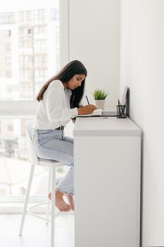 Young woman sitting at a desk writing, with natural light streaming through a window.