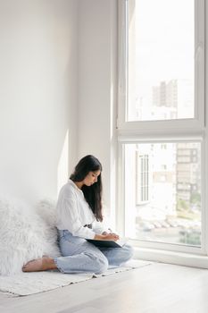 A woman sits indoors by a window, engaging in reading on a sunny day.