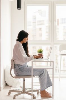 A young woman sits at a desk, typing on a laptop in a bright and cozy home setting.