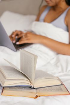 A person comfortably reading books and using a laptop in bed, draped in a white blanket.