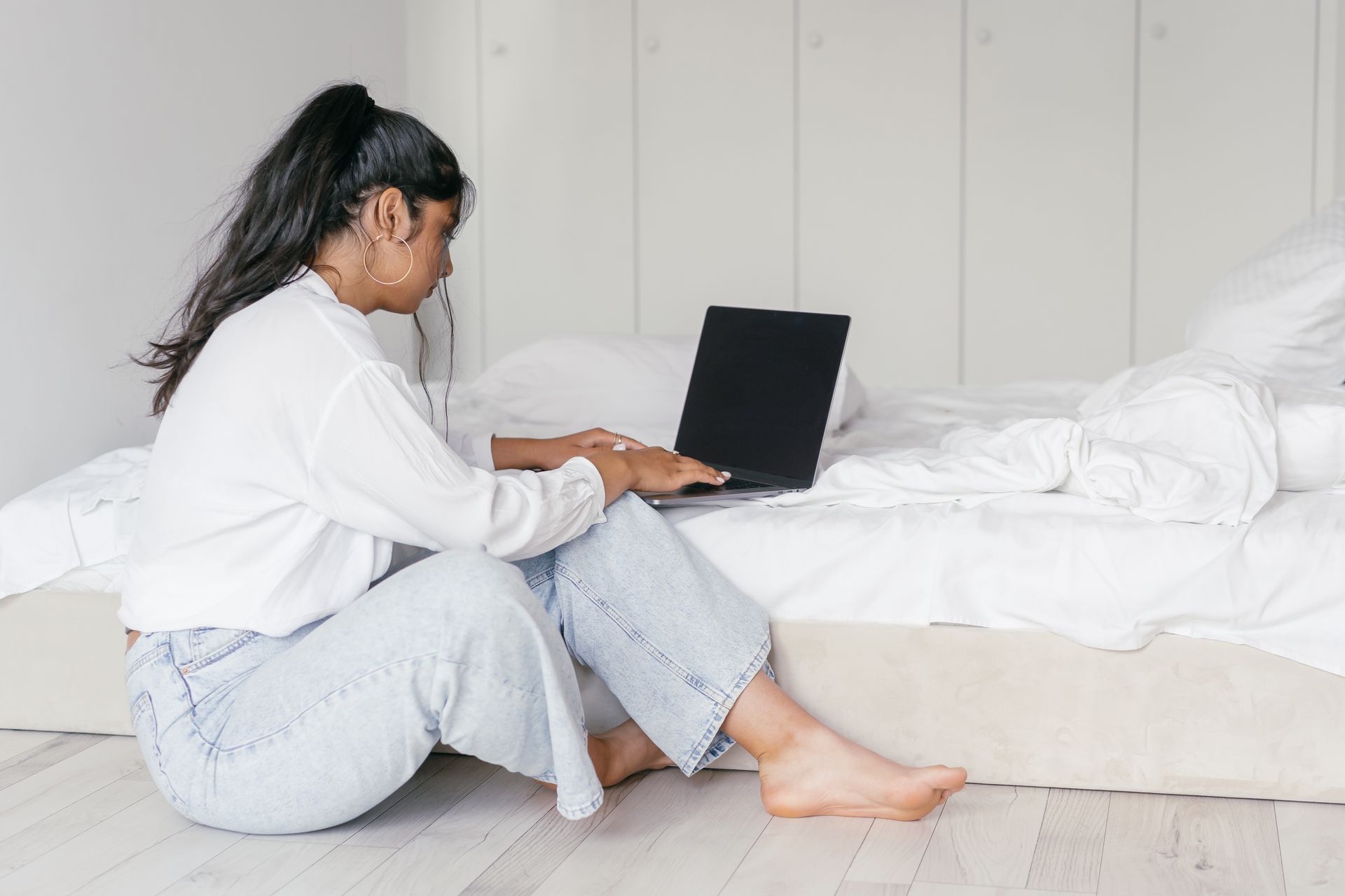 A woman sits on the floor working on a laptop in a serene bedroom environment, embodying remote work and relaxation.