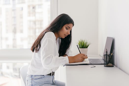 Young woman studying at a bright home desk with a laptop and notebook, focused on writing.