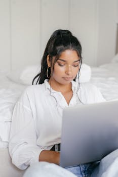 A young woman in a white shirt is focused on her laptop, sitting comfortably indoors.