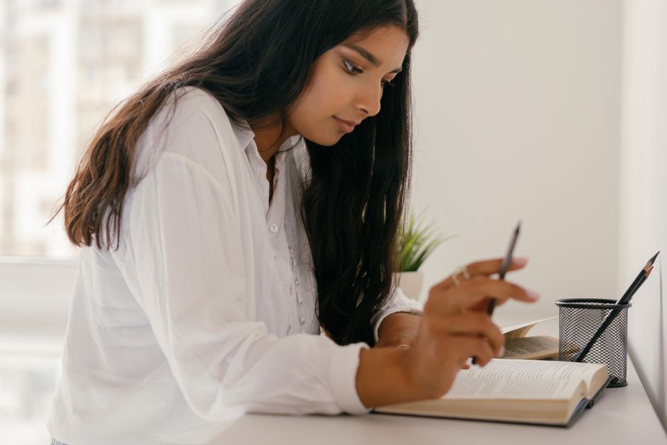 Young woman studying and writing notes at home, sitting by the window for work or homework. Young woman studying and writing notes at home, sitting by the window for work or homework.