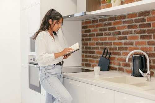 A Woman Standing Near the Kitchen Sink While Reading a Book