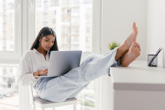 Woman in casual attire sitting indoors with feet up, working on a laptop by the window.