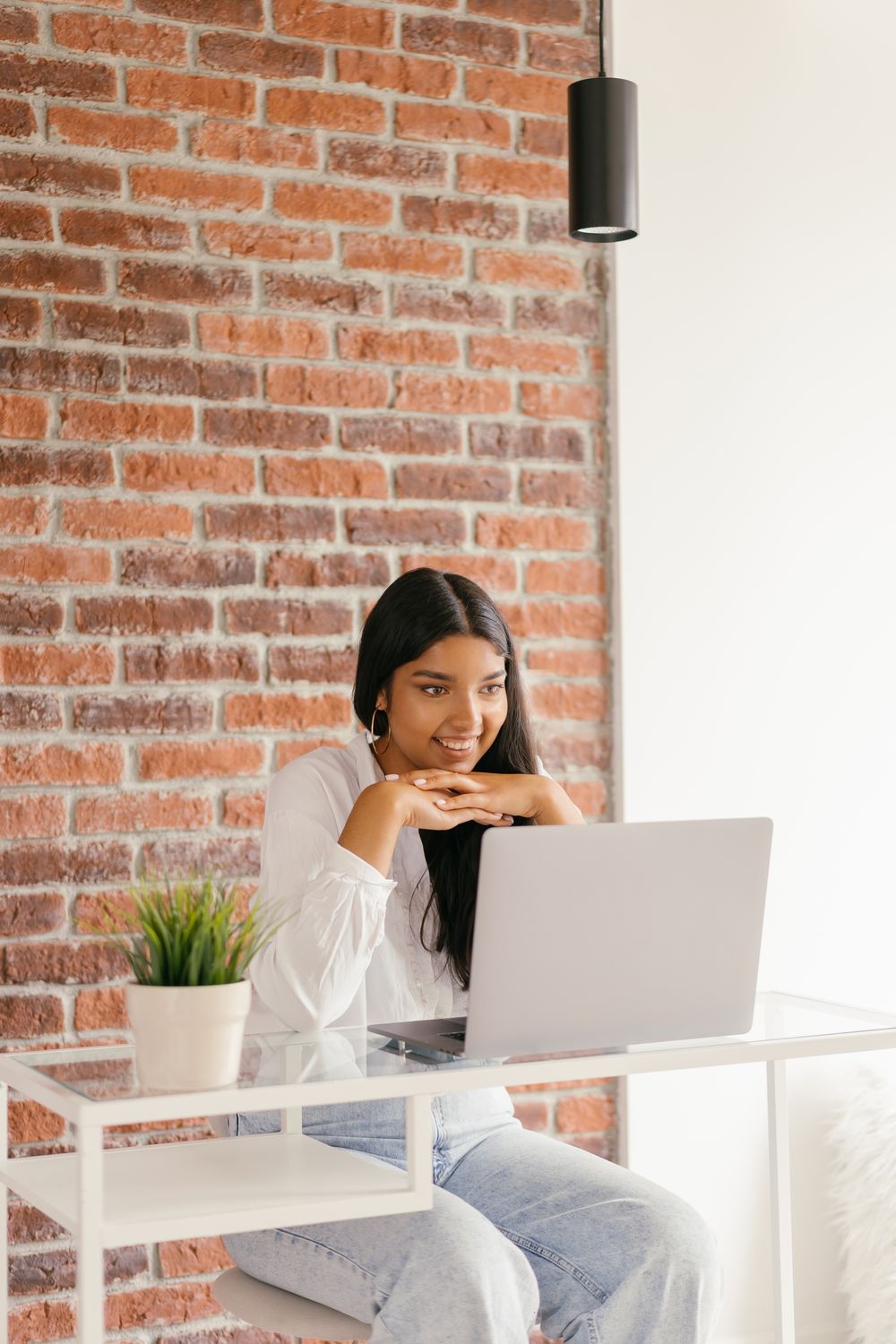 A woman rests her chin on her hands as she watches a lesson on her laptop at a glass desk in front of a red-brick wall.