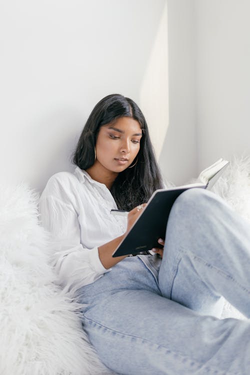 Woman in White Long Sleeve Shirt and Blue Denim Jeans Sitting on Bed Writing on Black Notebook