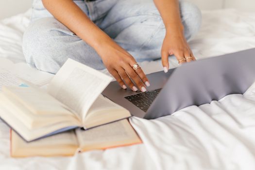 Woman multitasking between studying with books and typing on a laptop while seated on a bed.