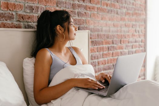 Woman using laptop in bed with brick wall backdrop, creating a cozy working environment.