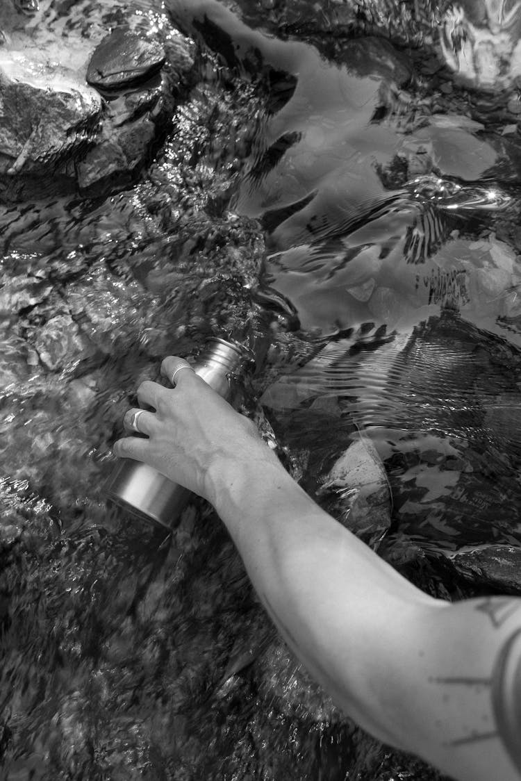 Grayscale Photo Of A Person Fetching Water On A Stream With Tumbler