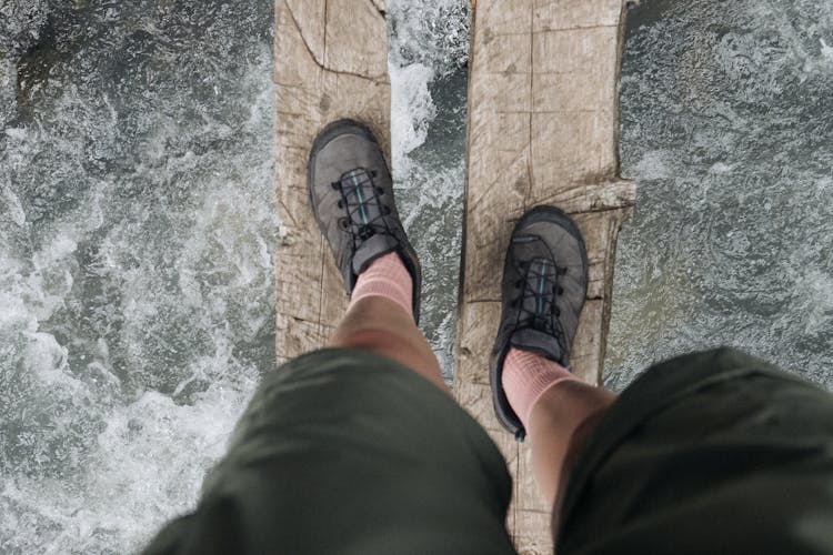 A Person Wearing Grey Hiking Shoes Standing On A Wooden Log Above A Flowing River