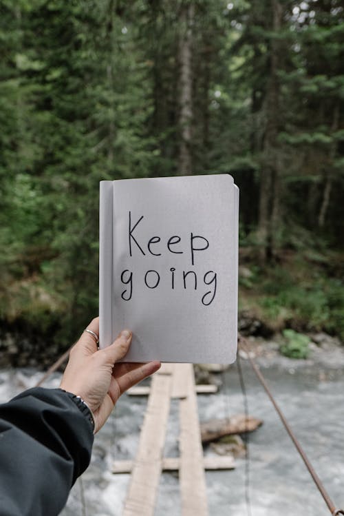 Person Holding a Notebook with a Text "Keep Going" on the Background of a River in a Forest 