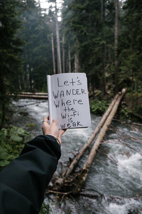 Person Holding a Notebook with a Text on the Background of a River in a Forest 