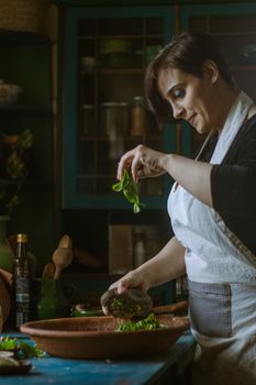 A woman in a kitchen prepares a fresh green salad, adding herbs with care and precision.