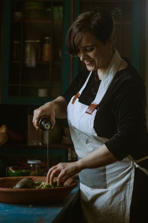 Free Woman in kitchen pouring olive oil over a dish, wearing apron, mid-preparation. Stock Photo