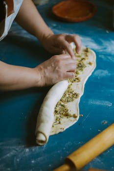 Close-up of hands rolling herb-filled dough on a blue table, capturing culinary artistry.