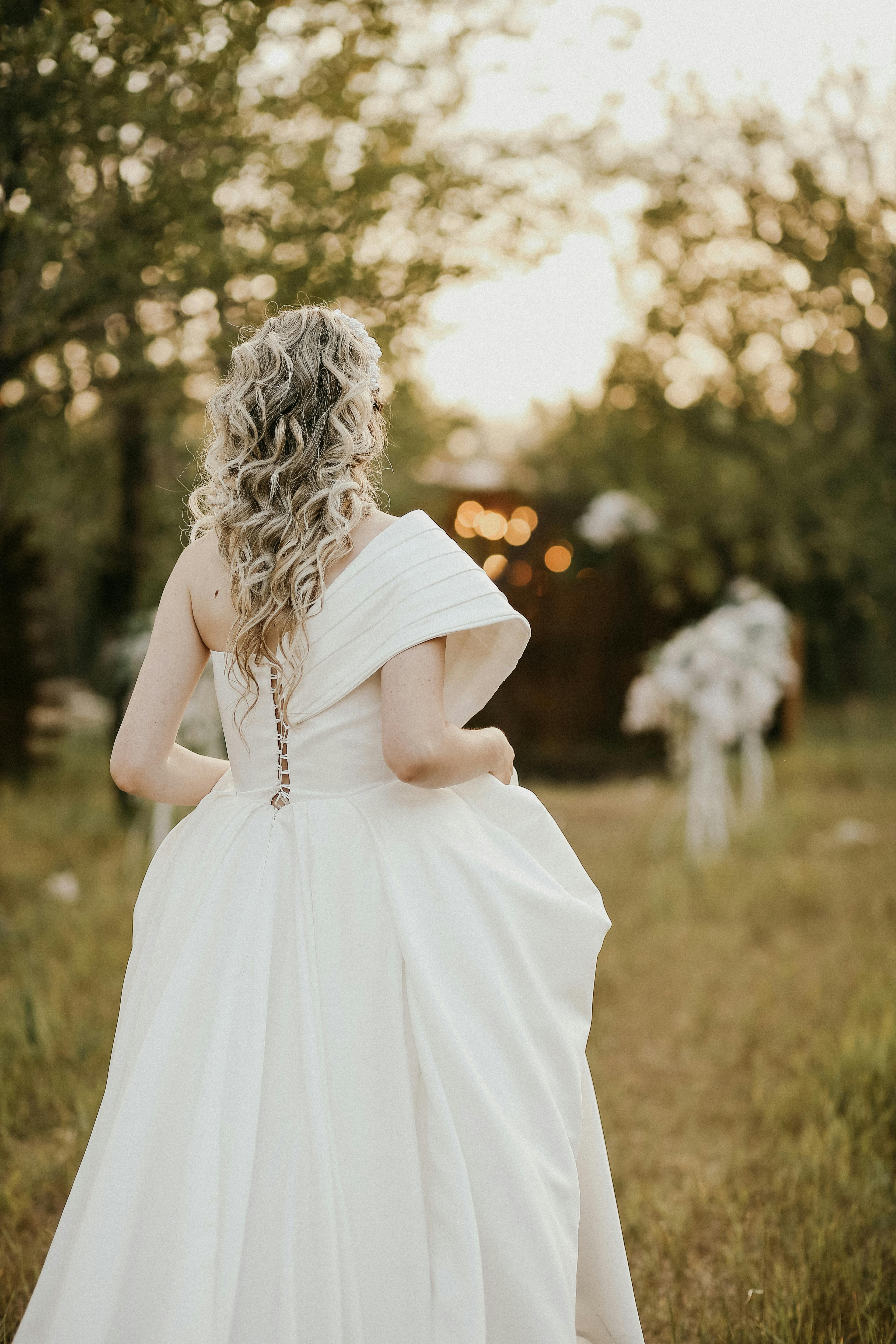 A bride with blonde hair in a white gown walking in a serene outdoor wedding setting.