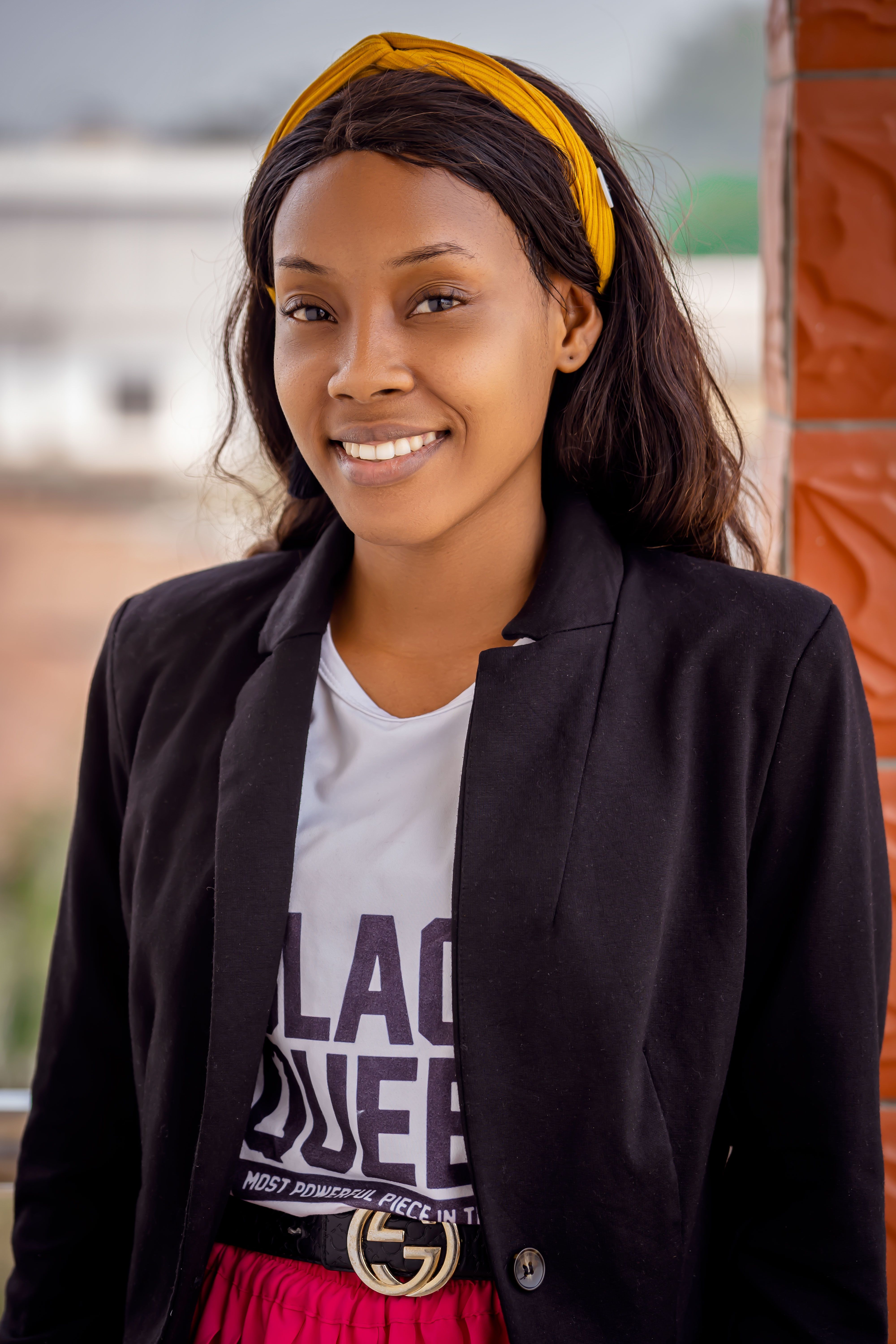 Portrait of a young woman smiling warmly, wearing a black blazer and yellow headband, outdoors.