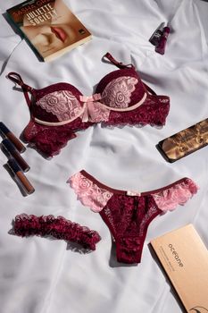 Sophisticated burgundy and pink lace lingerie set arranged with makeup and books for a stylish flat lay.