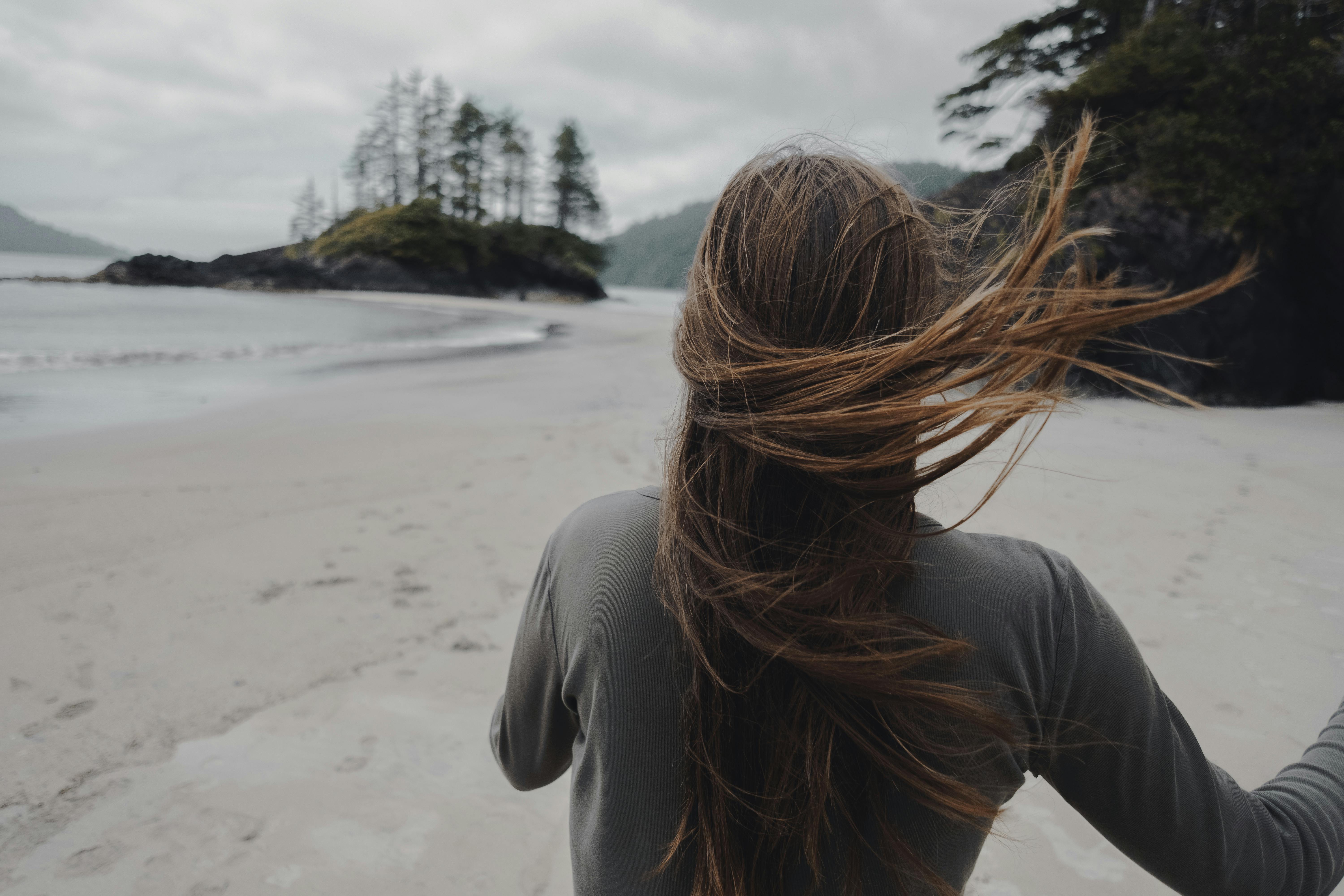 A woman with long hair stands on a windy Tofino beach in British Columbia, overlooking the scenic coastline.
