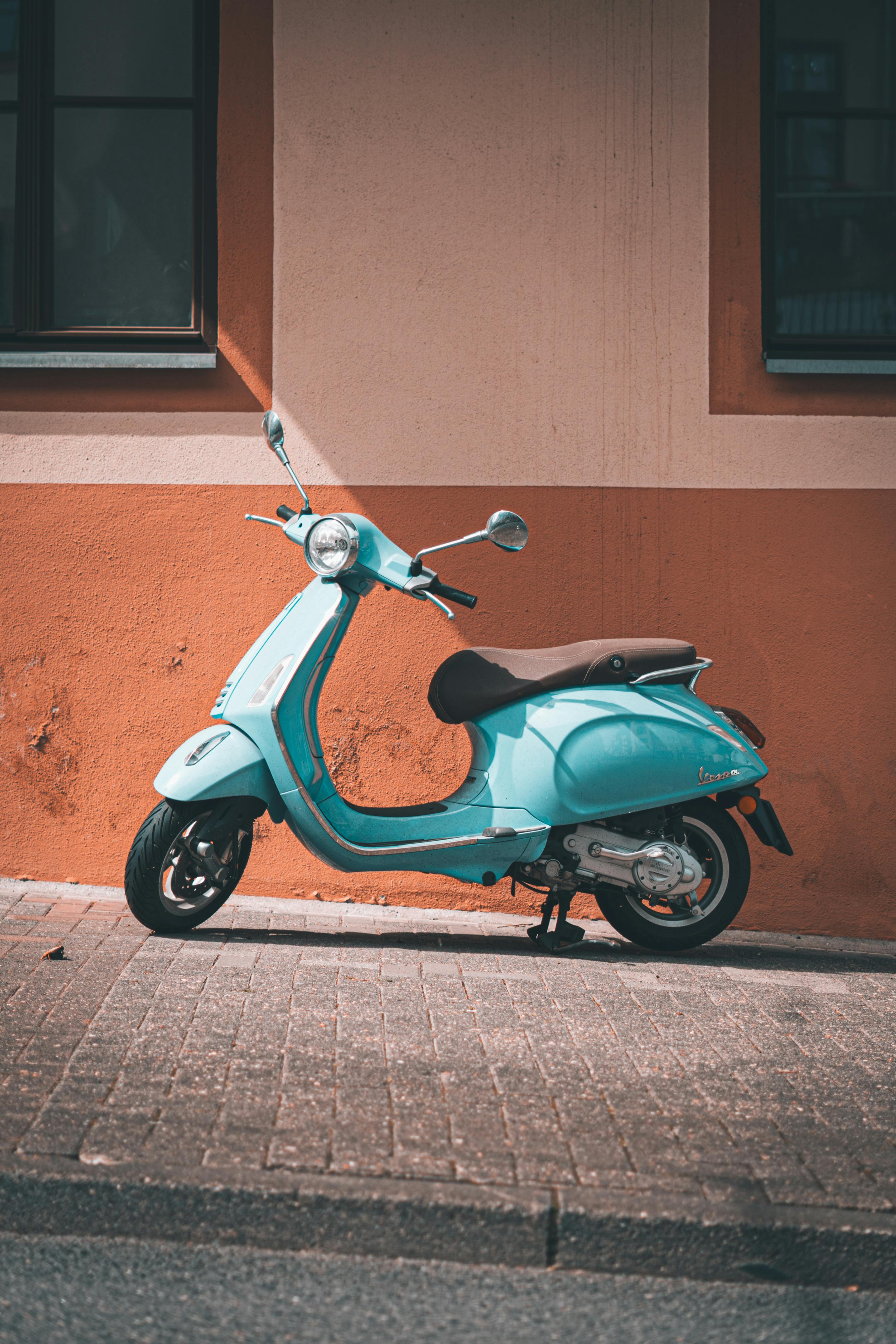 Free A classic blue Vespa scooter parked against a rustic orange wall on a cobblestone street. Stock Photo