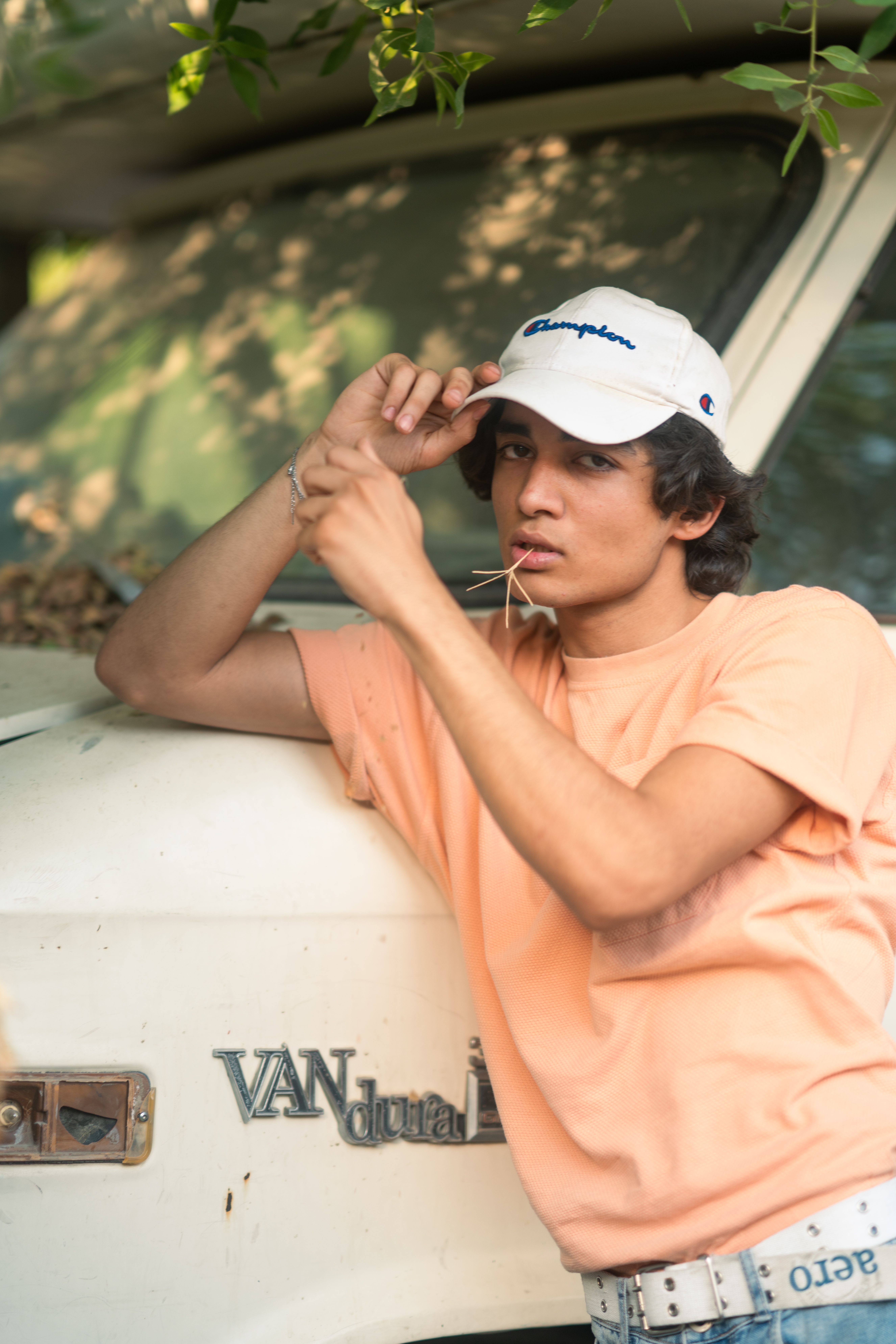 A relaxed young man poses against a classic van outdoors wearing a cap and casual attire.