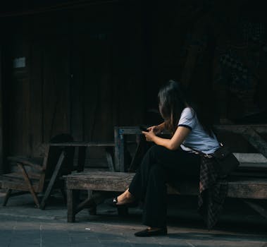 A young woman in casual attire is focused on her phone while sitting on a rustic wooden bench.